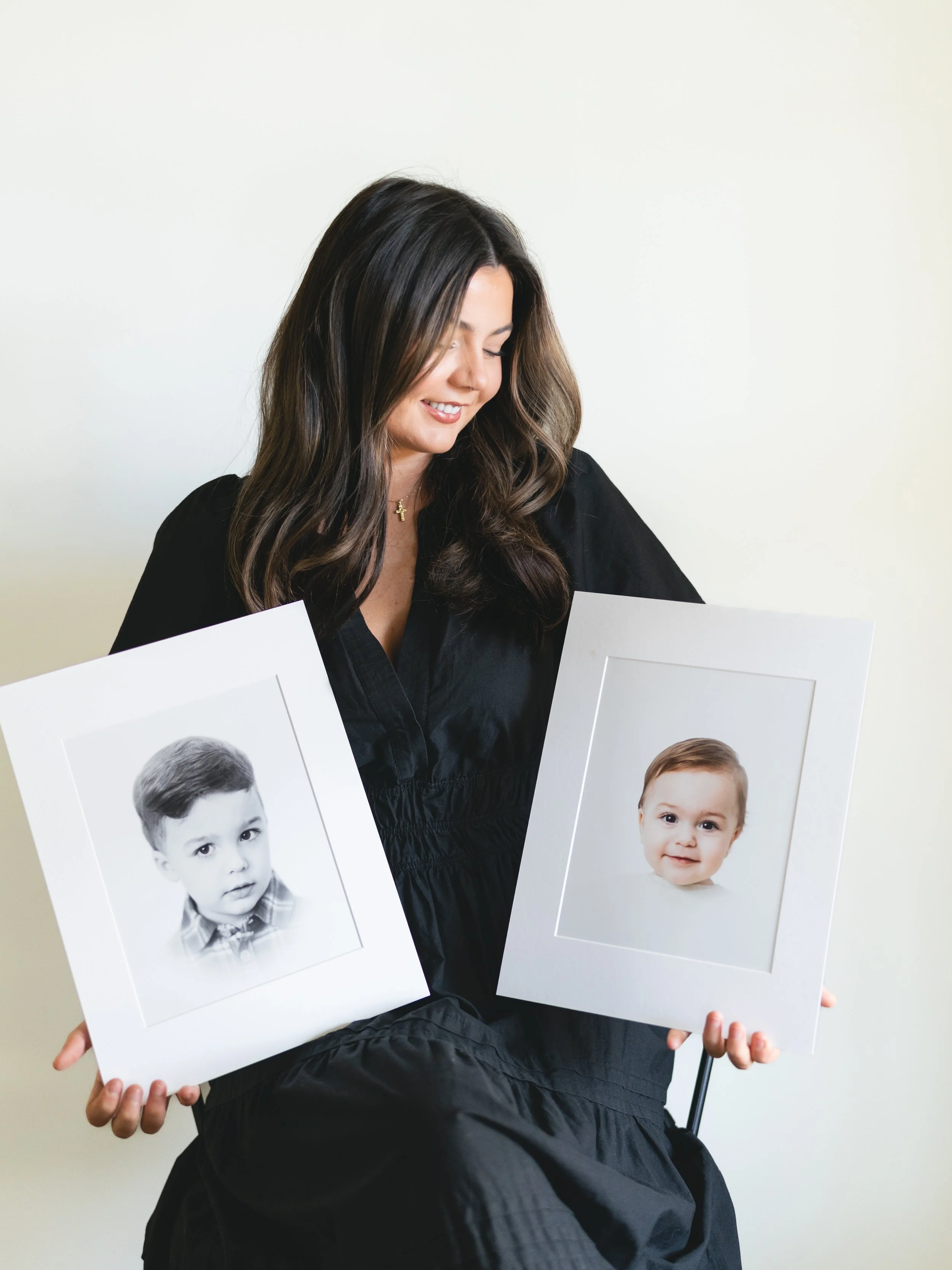 A woman with long dark hair is smiling and looking down, holding two framed photos of children, one black and white and one in color, against a plain light background.