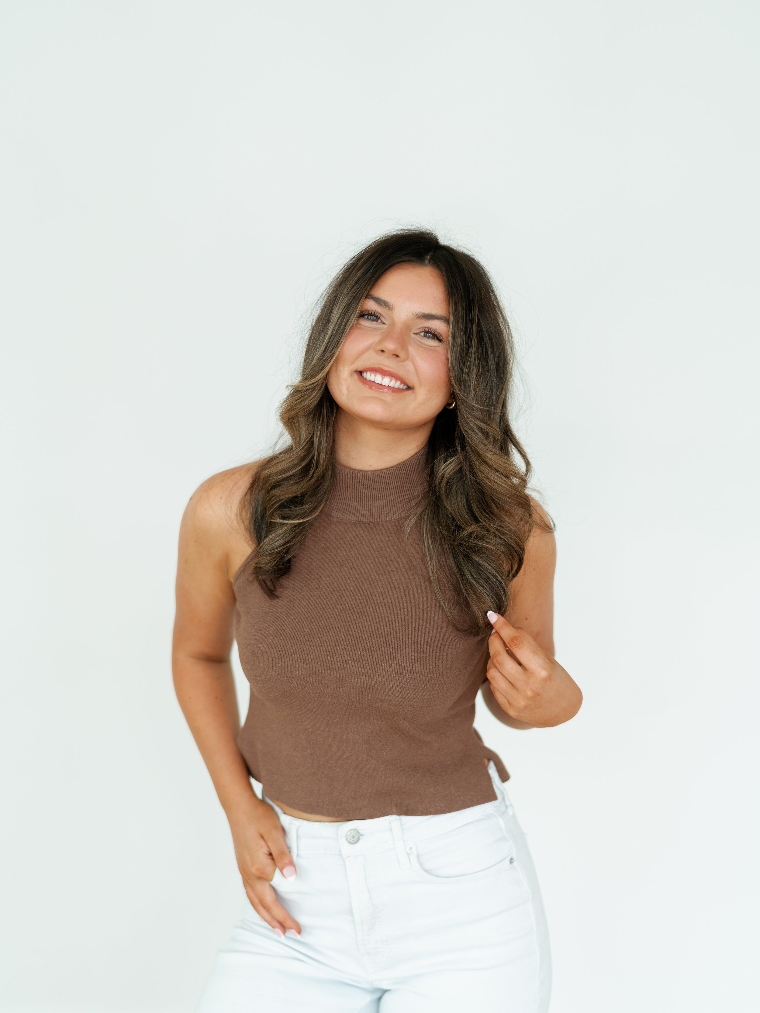 A woman with long wavy brown hair, wearing a sleeveless brown top and white jeans, smiling, pointing to the side with her right hand, against a plain white background.