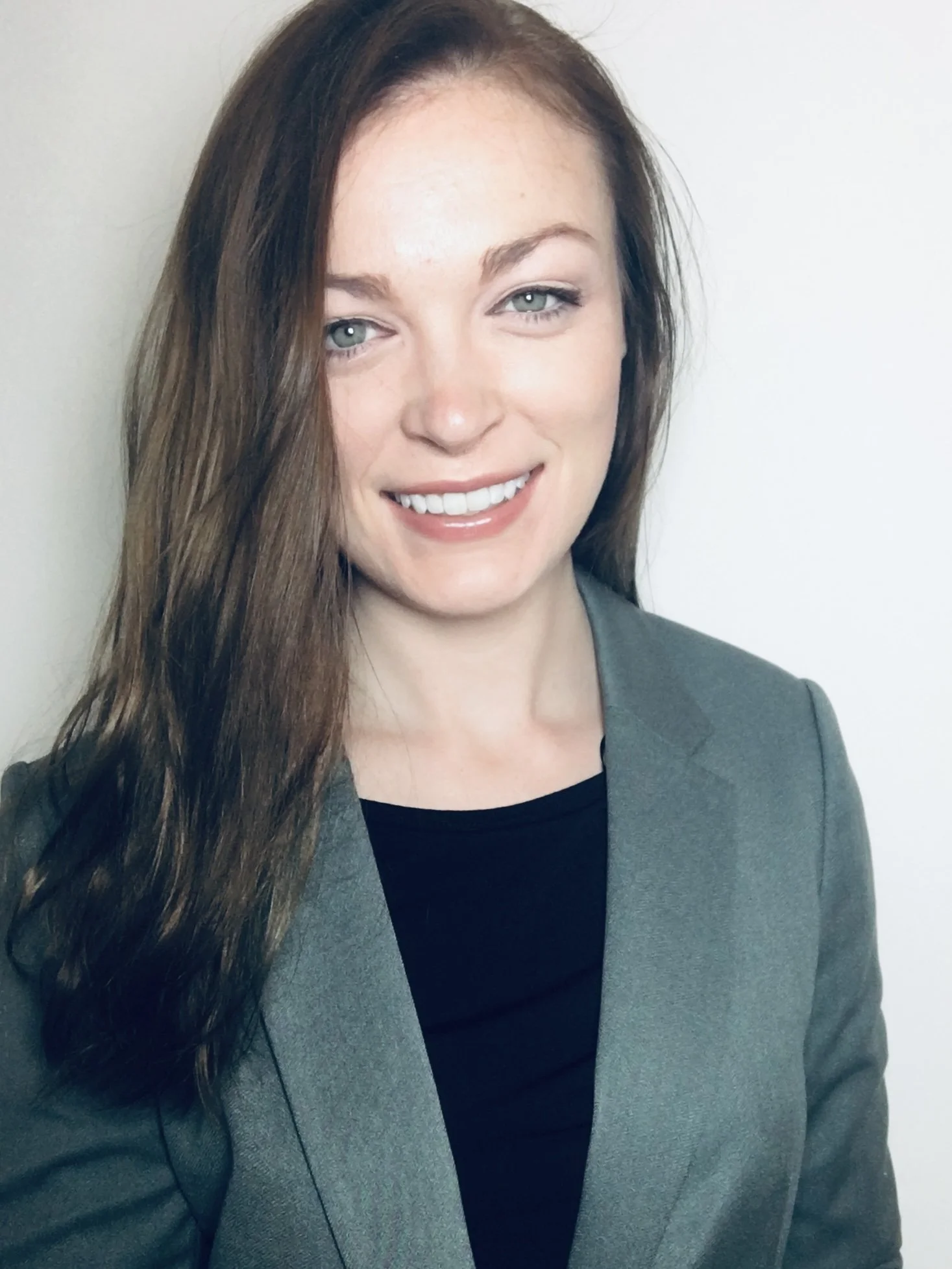 Smiling woman with long brown hair, green eyes, wearing a gray blazer and black top, standing against a plain white background.