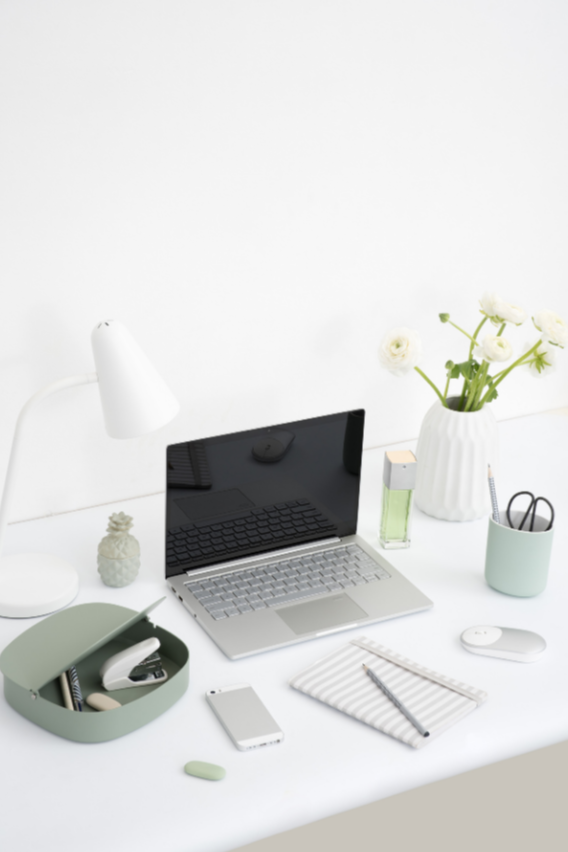 Minimalist white desk with a silver laptop, white mouse, and smartphone, accompanied by office supplies, a green container, a white vase with white roses, a clear perfume bottle, a pencil holder, and a wireless drawer on a white wall background.