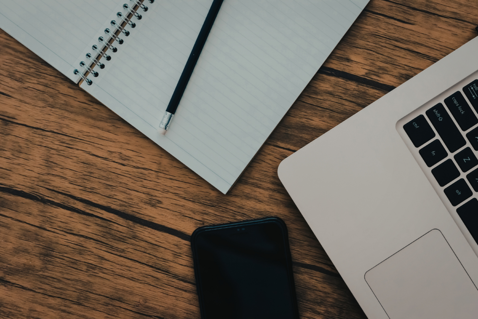 A wooden desk with an open laptop, a black phone, a spiral notebook, and a pencil placed on top of the notebook.