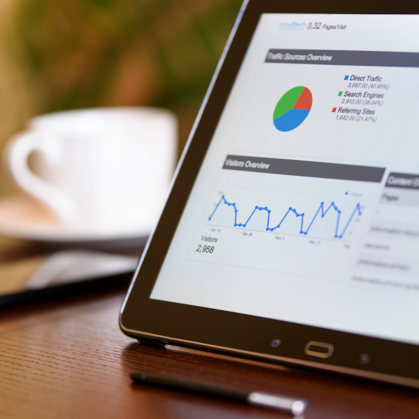 Tablet displaying website analytics including traffic sources and visitor overview, placed on a wood table with a pen nearby and a blurred coffee mug in the background.
