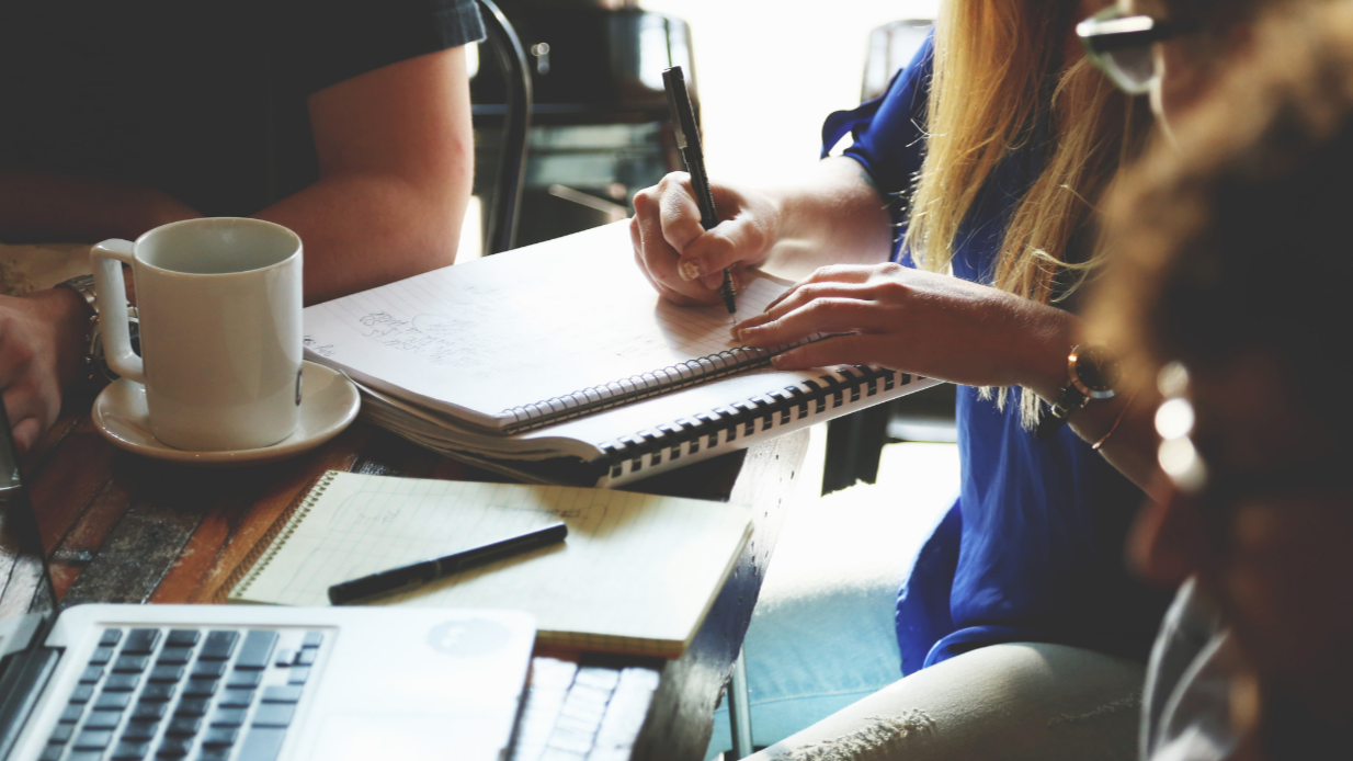 Three people working at a cluttered table with notebooks, a laptop, and a coffee mug.