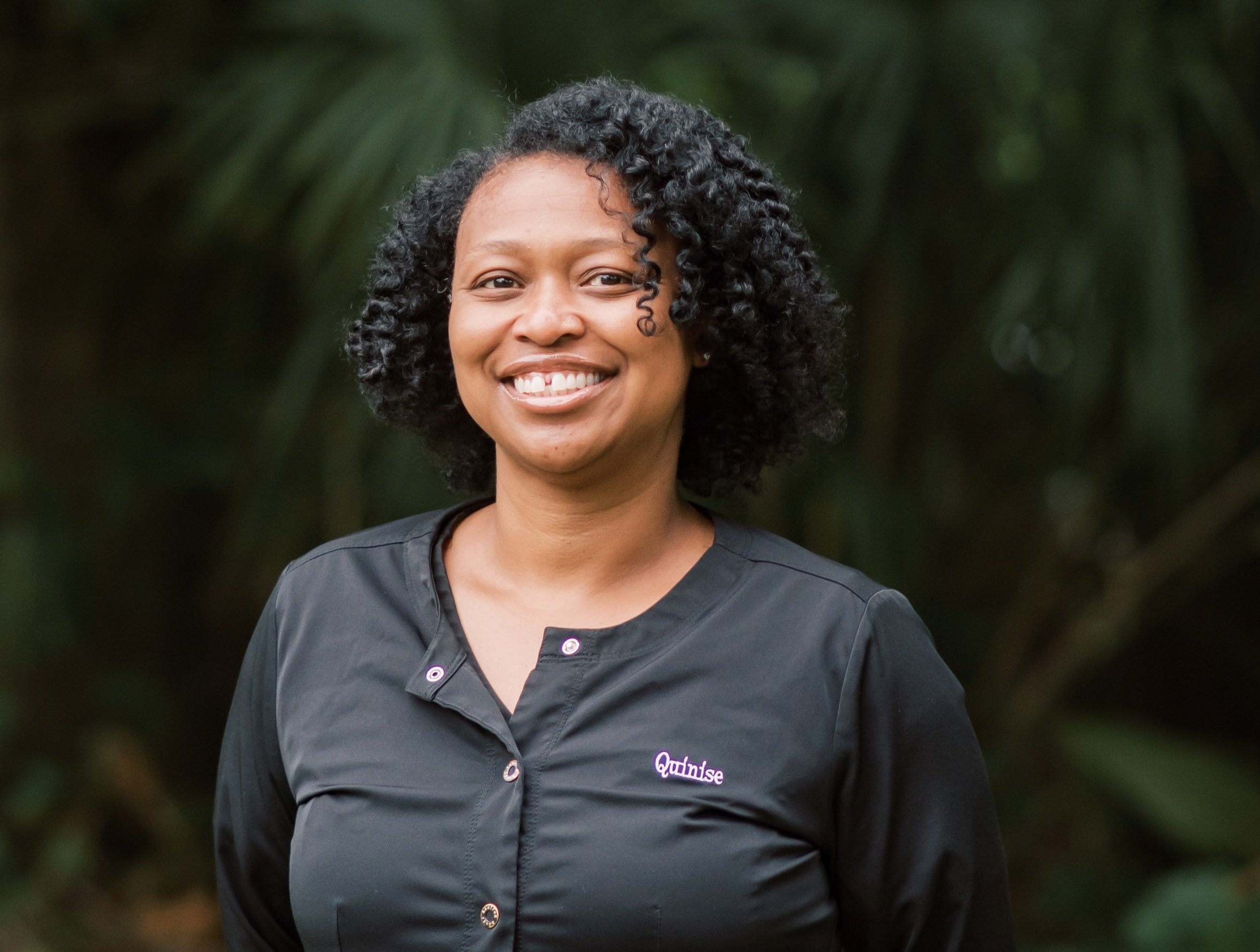 A woman with curly black hair smiling, wearing a dark gray shirt with the name 'Quintise' embroidered on it, standing outdoors with blurred green foliage in the background.