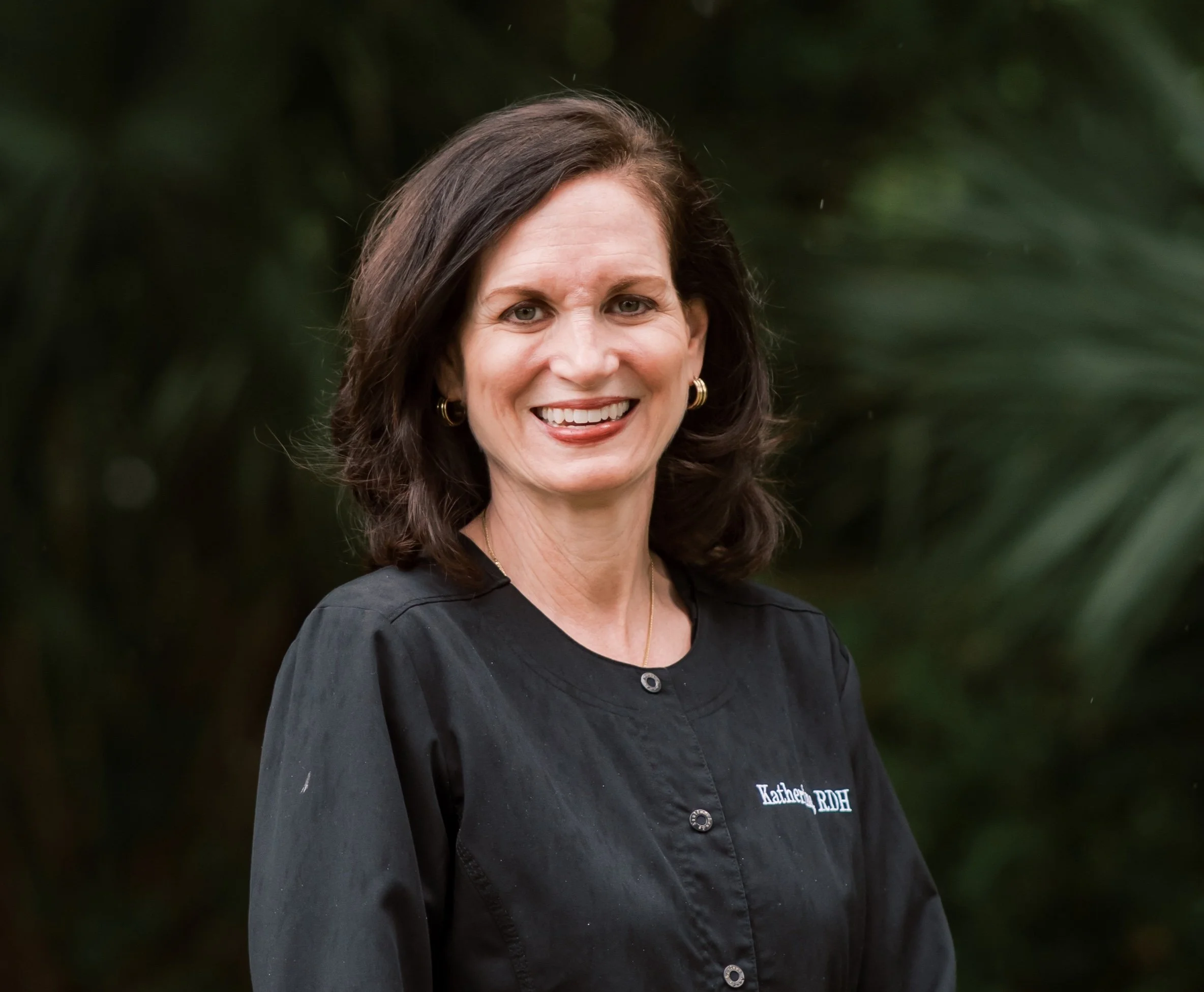A woman with dark brown hair, wearing a black shirt with embroidered text, smiling outdoors with a blurred green background.