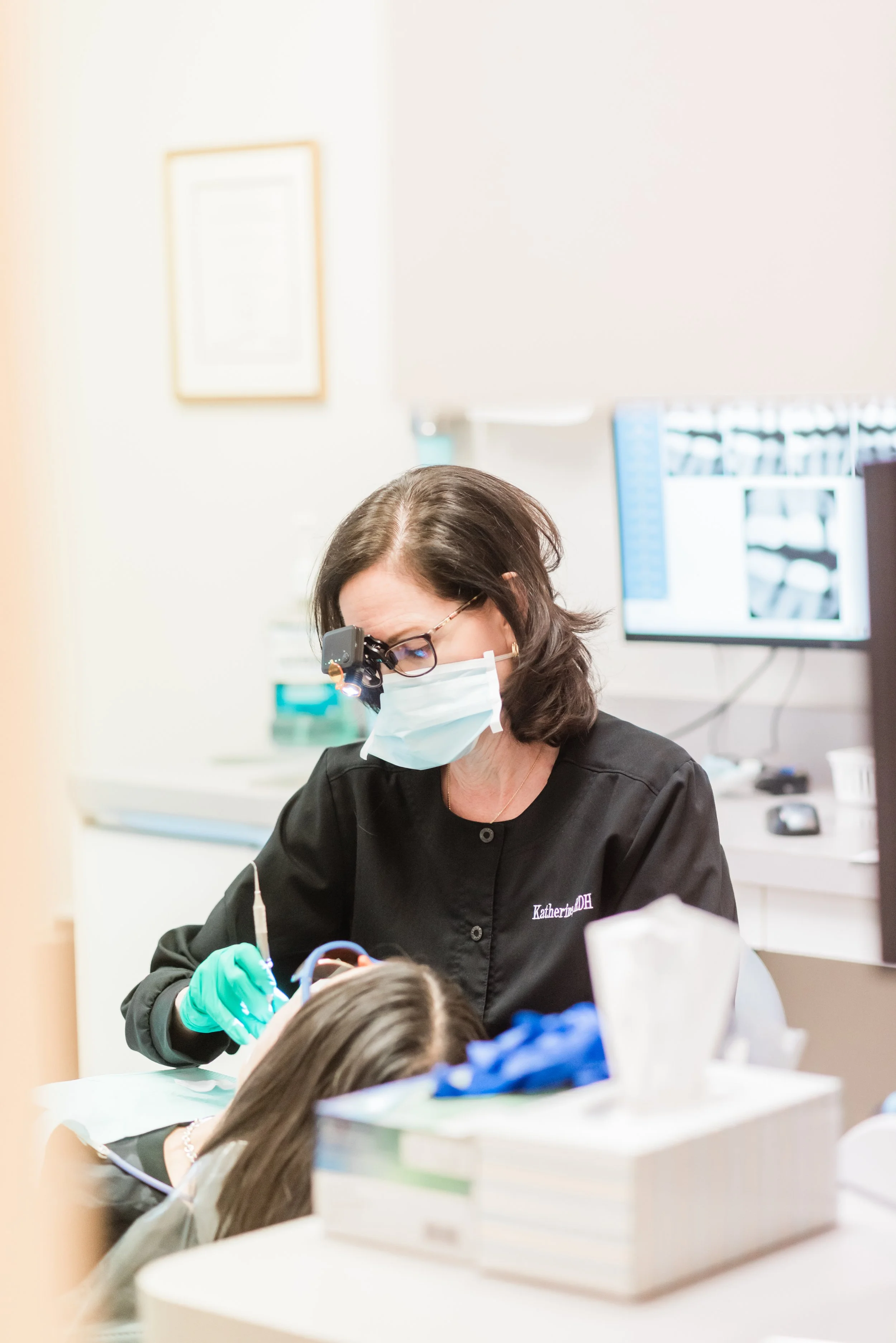 Dental professional wearing glasses, mask, and gloves providing dental care to a patient in a clinical setting, with dental equipment and monitors in the background.