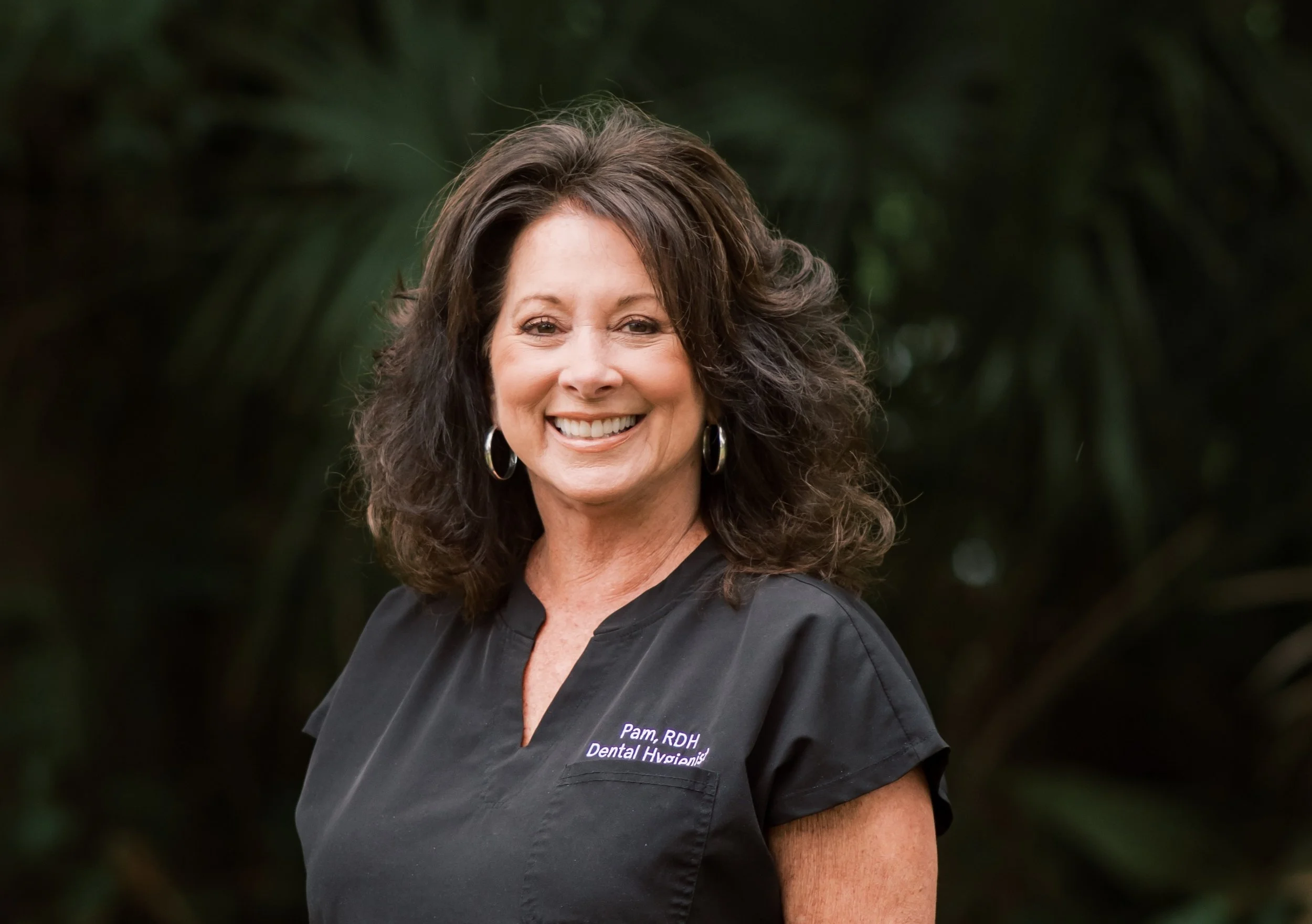 A smiling woman with dark, curly, shoulder-length hair wearing a black dental hygienist uniform with text that reads 'Pam, RDH Dental Hygienist', standing outdoors with a blurred green foliage background.