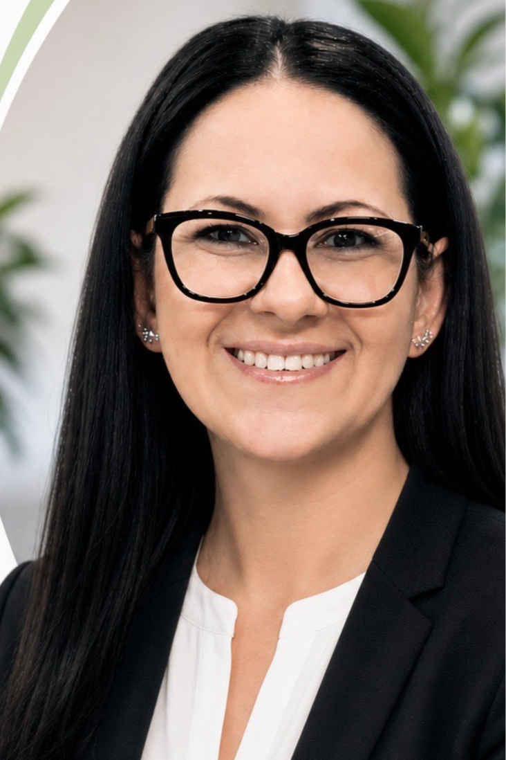 A woman with long black hair, wearing glasses, a white blouse, and a black blazer, smiling at the camera with a blurred background of green plants.