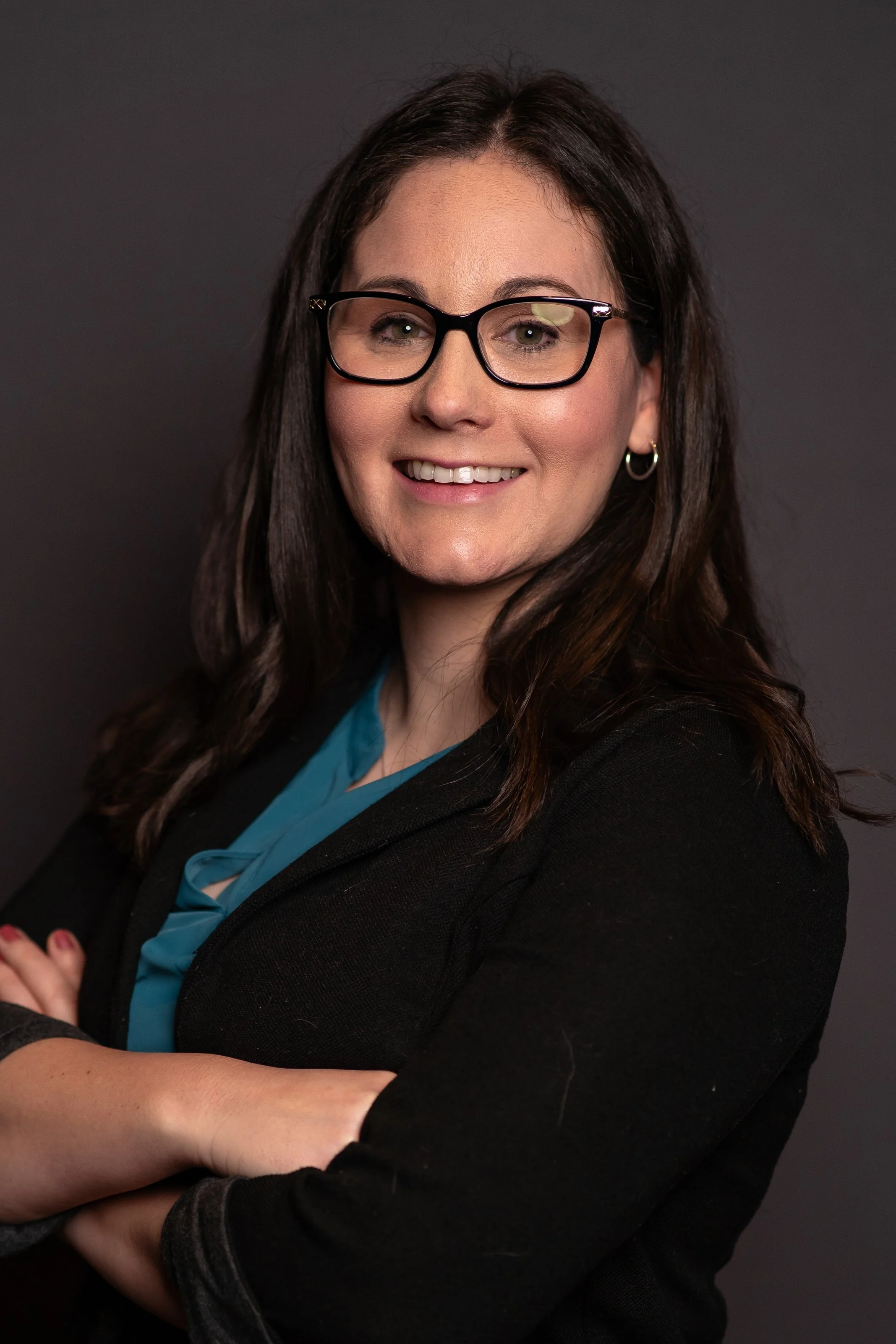 A woman with long dark hair, glasses, and earrings, smiling with her arms crossed against a dark background.