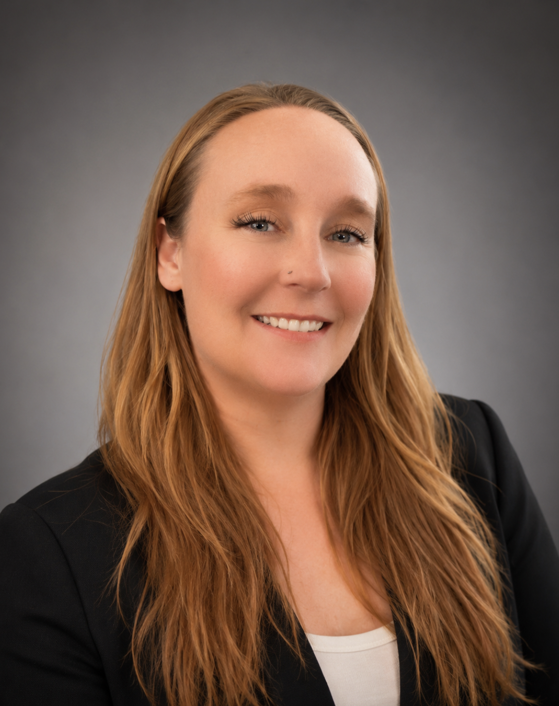 A woman with long red hair and light makeup, wearing a black blazer and a white top, smiling against a gray background.