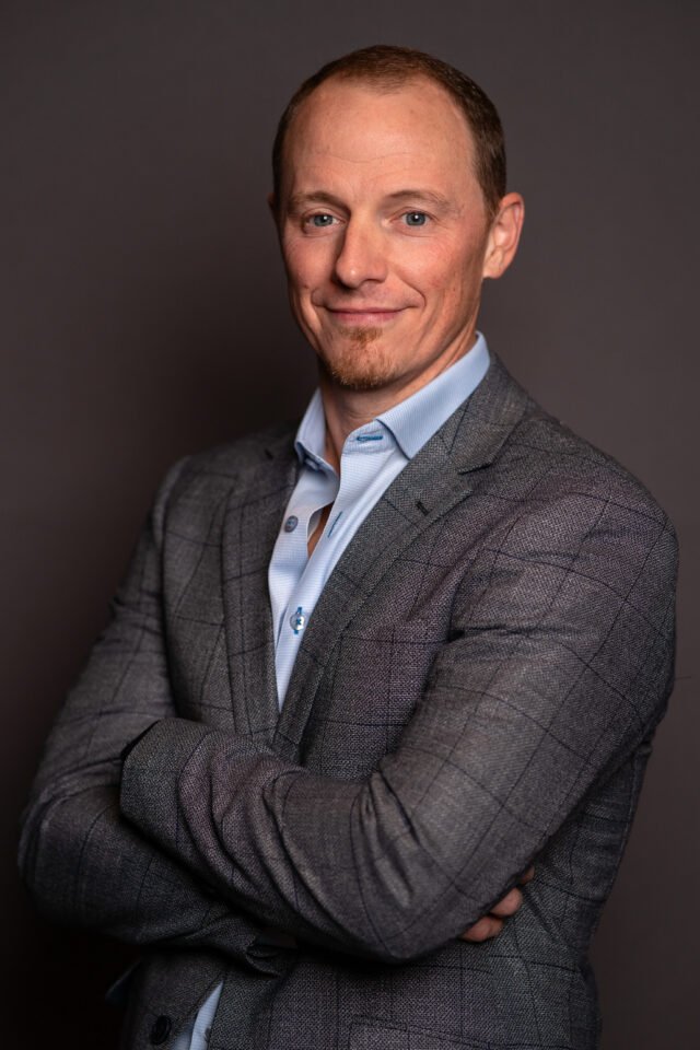 A professional headshot of a man in a gray checked blazer and light blue shirt, posing with arms crossed against a dark gray background.
