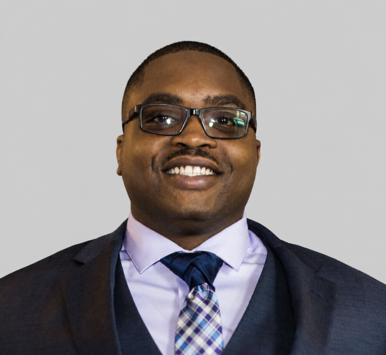 A smiling man in a suit with glasses and a patterned tie, posing against a neutral background.