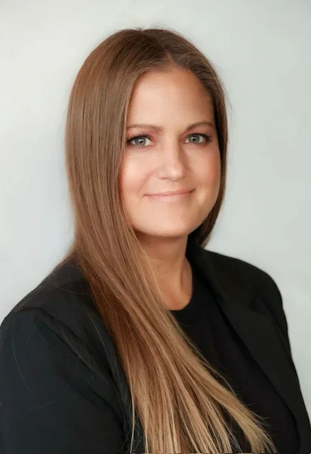 A woman with long, straight, light brown hair, light skin, and blue eyes, wearing a black top and blazer, smiling softly against a light gray background.