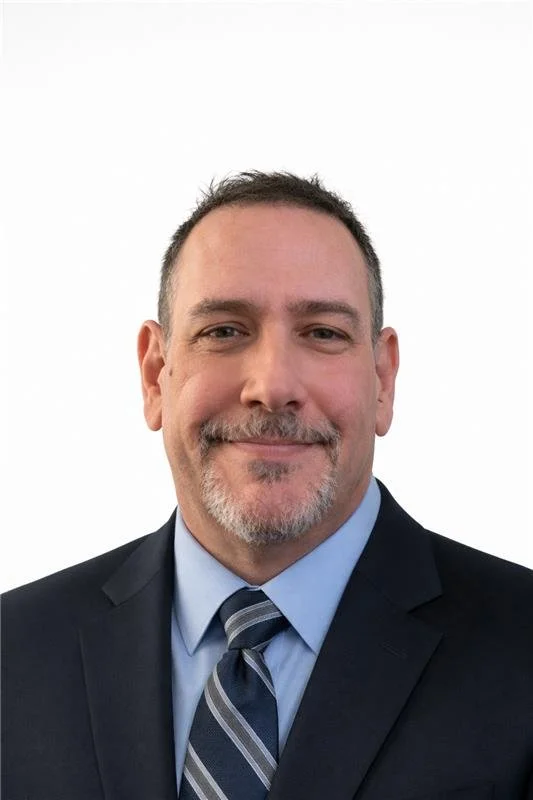 A professional headshot of a middle-aged man with short dark hair, a trimmed beard, wearing a dark suit, light blue dress shirt, and a striped tie, smiling in front of a plain white background.