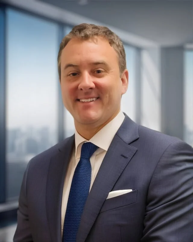 A smiling man in a navy blue suit, white shirt, and blue tie, standing in an office with large windows and a city view in the background.