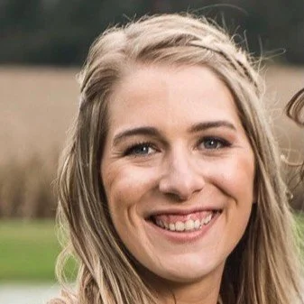 A smiling young woman with long blond hair outdoors in a natural setting.