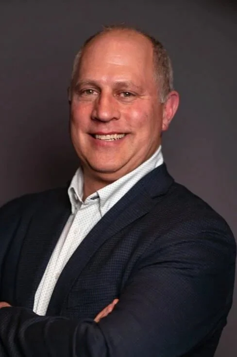 A smiling man in a dark blazer and white shirt with arms crossed, standing against a dark gray background.