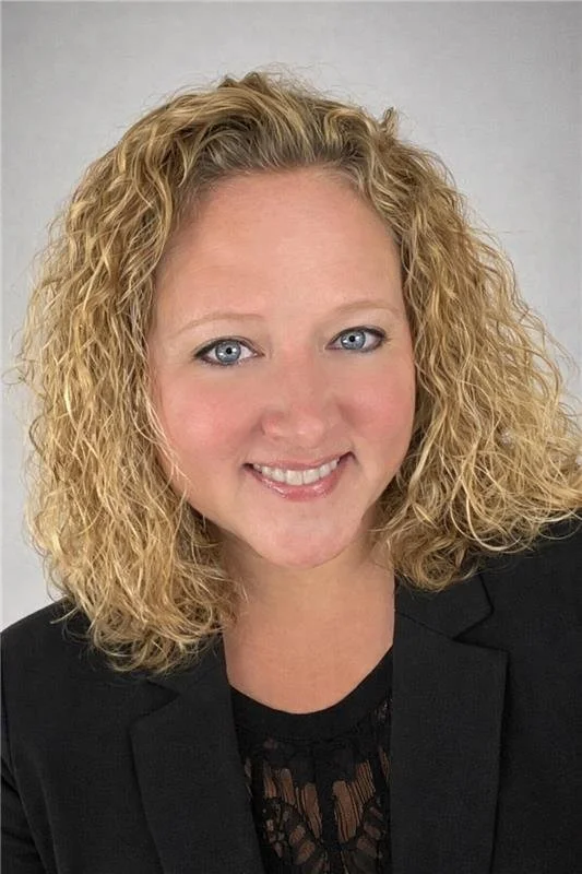 A woman with shoulder-length curly blonde hair, blue eyes, and fair skin, smiling at the camera, wearing a black blazer over a black lace top, against a plain light gray background.