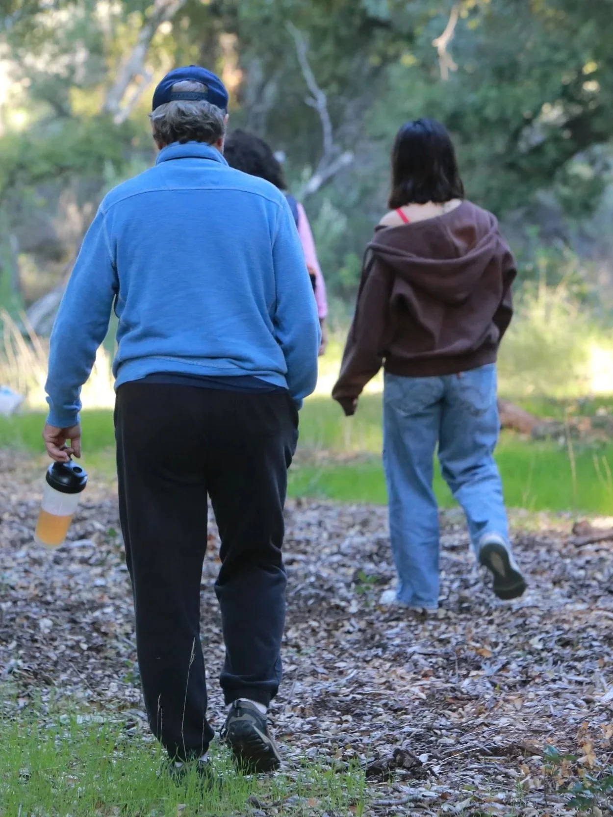 Three people hiking on a wooded trail, viewed from behind, with one person holding a water bottle.