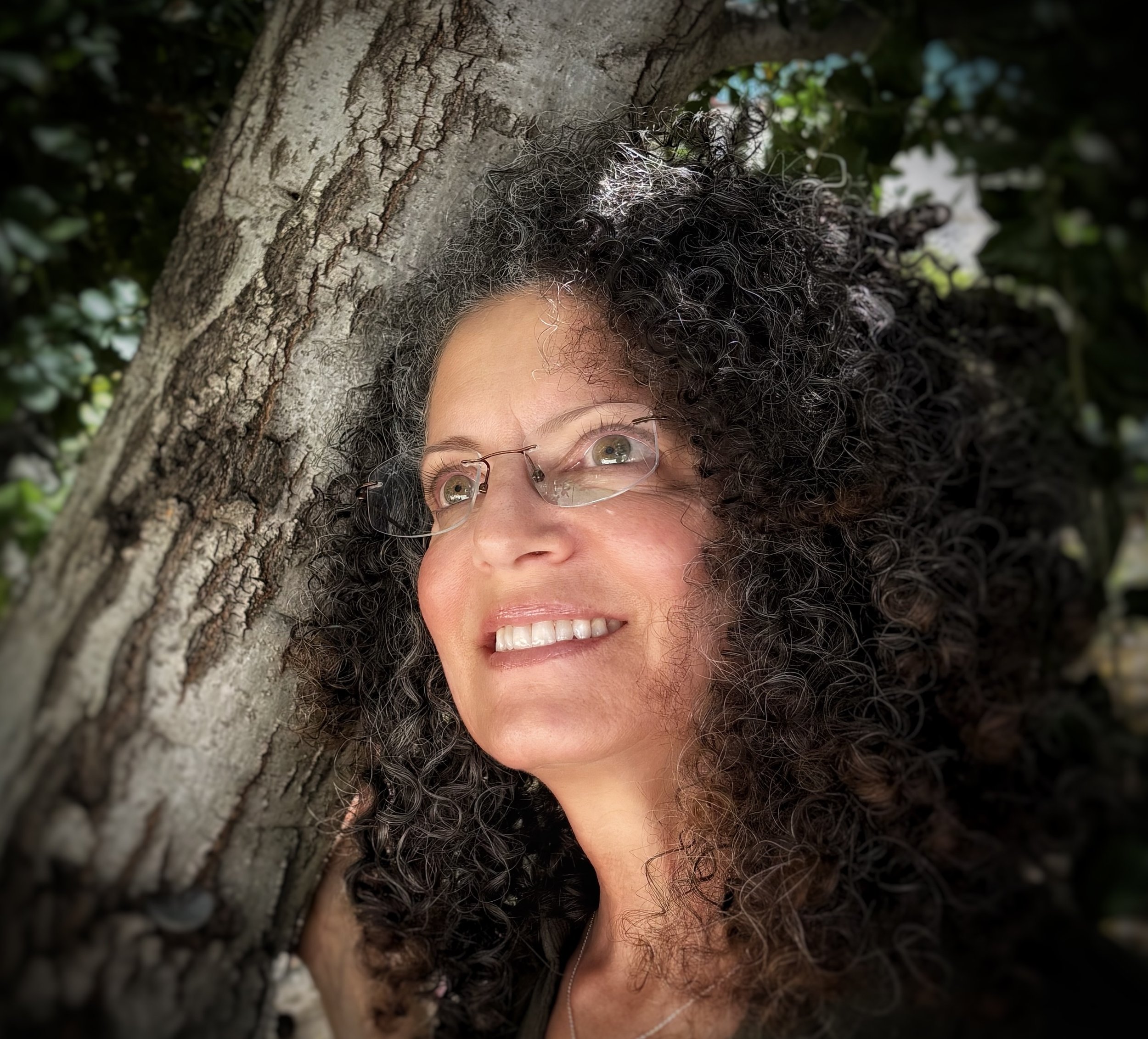 Woman with curly dark hair and glasses leaning against a tree, smiling, with green foliage in the background.