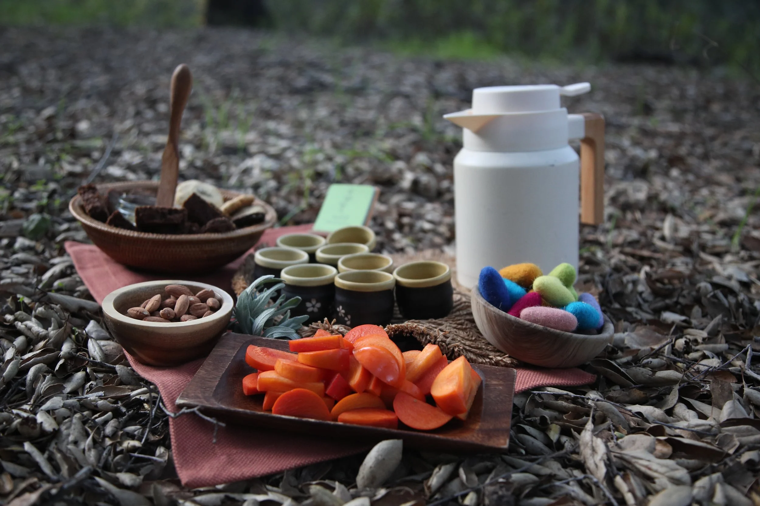 A picnic setup on a bed of dry leaves with various snacks and items, including a wooden platter of sliced carrots, a bowl of mixed nuts, a basket of colorful felt balls, small cups, a white bottle, and bowls of snacks, with a pink cloth underneath.