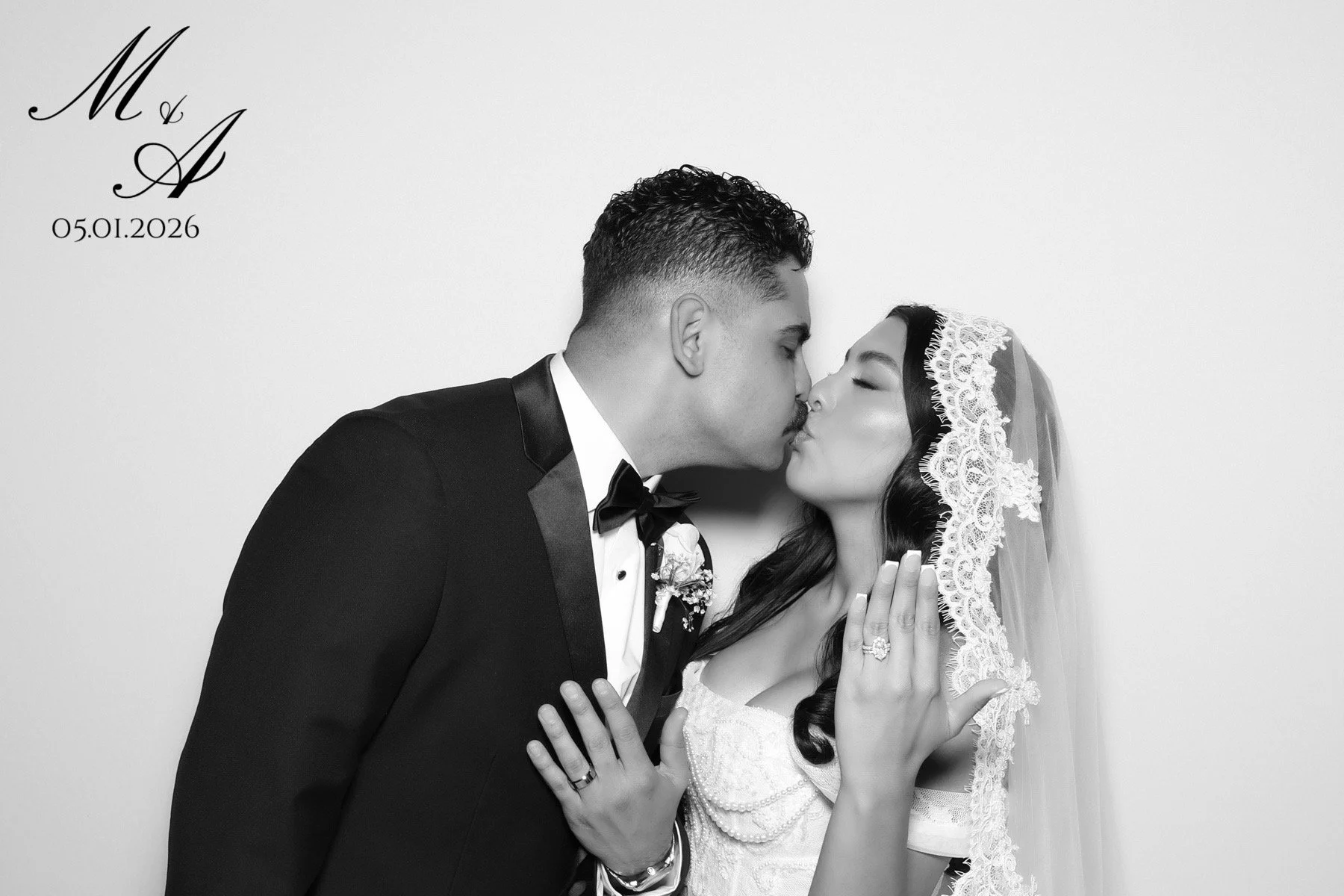 A black-and-white photo of a groom in a tuxedo and a bride in a wedding dress sharing a kiss. The bride wears a lace veil, and the groom wears a boutonniere. The bride has her hands on the groom's chest, showing her wedding ring. The upper left corner has initials "M & A" and the date "05.01.2026."