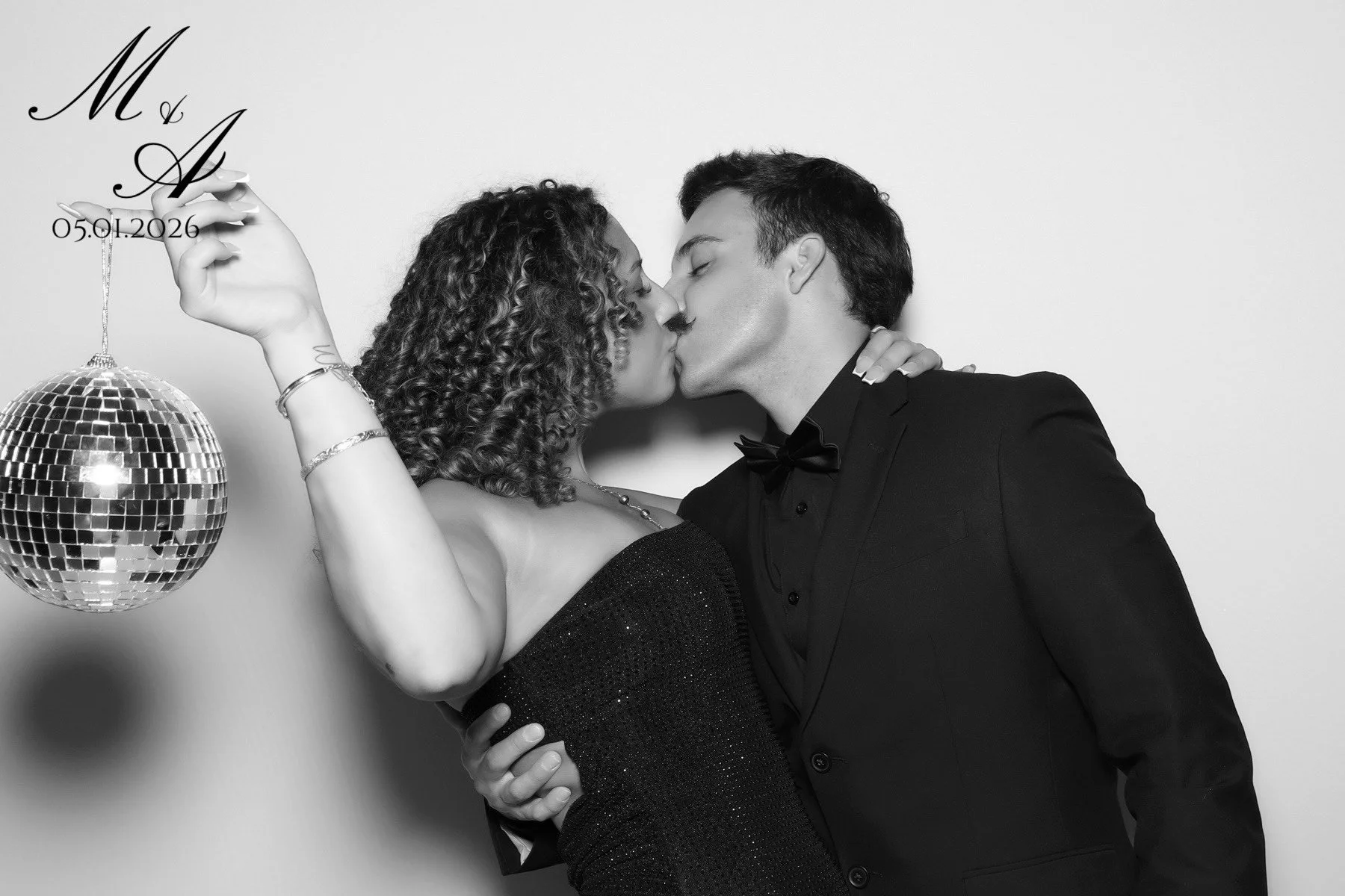 A black and white photo of a couple kissing; the woman has curly hair and is wearing a black dress, while the man is in a tuxedo with a bow tie. The woman is holding a disco ball and a string with lettering and a date, and their arms are around each other against a plain background.