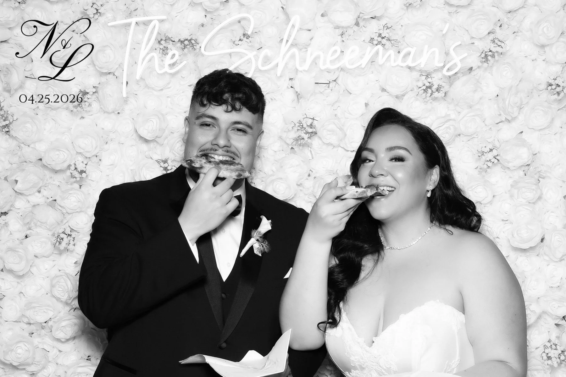 Black and white photo of a bride and groom in wedding attire, smiling and holding slices of pizza, standing against a floral backdrop with the words 'The Schneemans' and date '04.25.2026' in neon-style lettering.