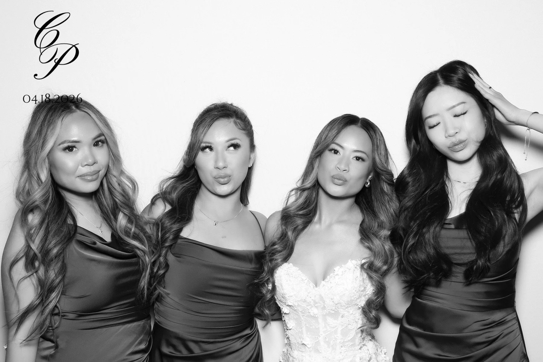 Four women in formal dresses, posing against a plain background. One woman in the middle is wearing a white lace wedding dress, while the others are in dark satin dresses. All have long, styled hair and are looking at the camera or in different directions.