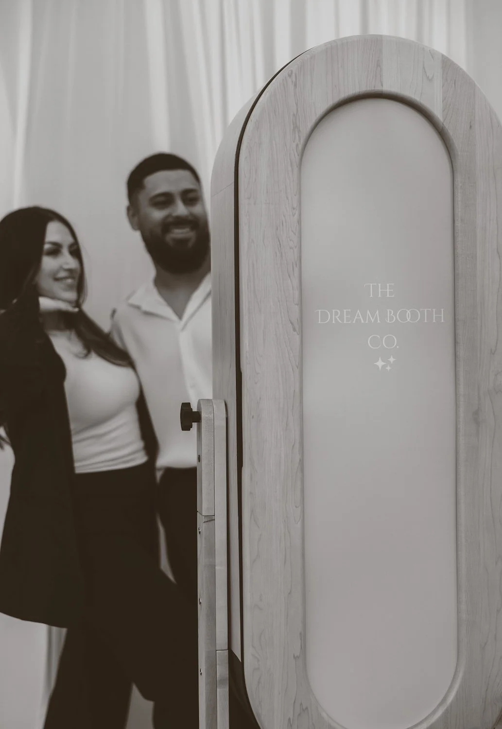 Melissa and Kevin Najarro stand next to a photo booth labeled 'The Dream Booth Co.' in a well-lit room with white curtains.
