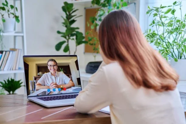 Woman participating in a video call with a woman on a laptop screen, sitting at a wooden table in a bright room with green plants.