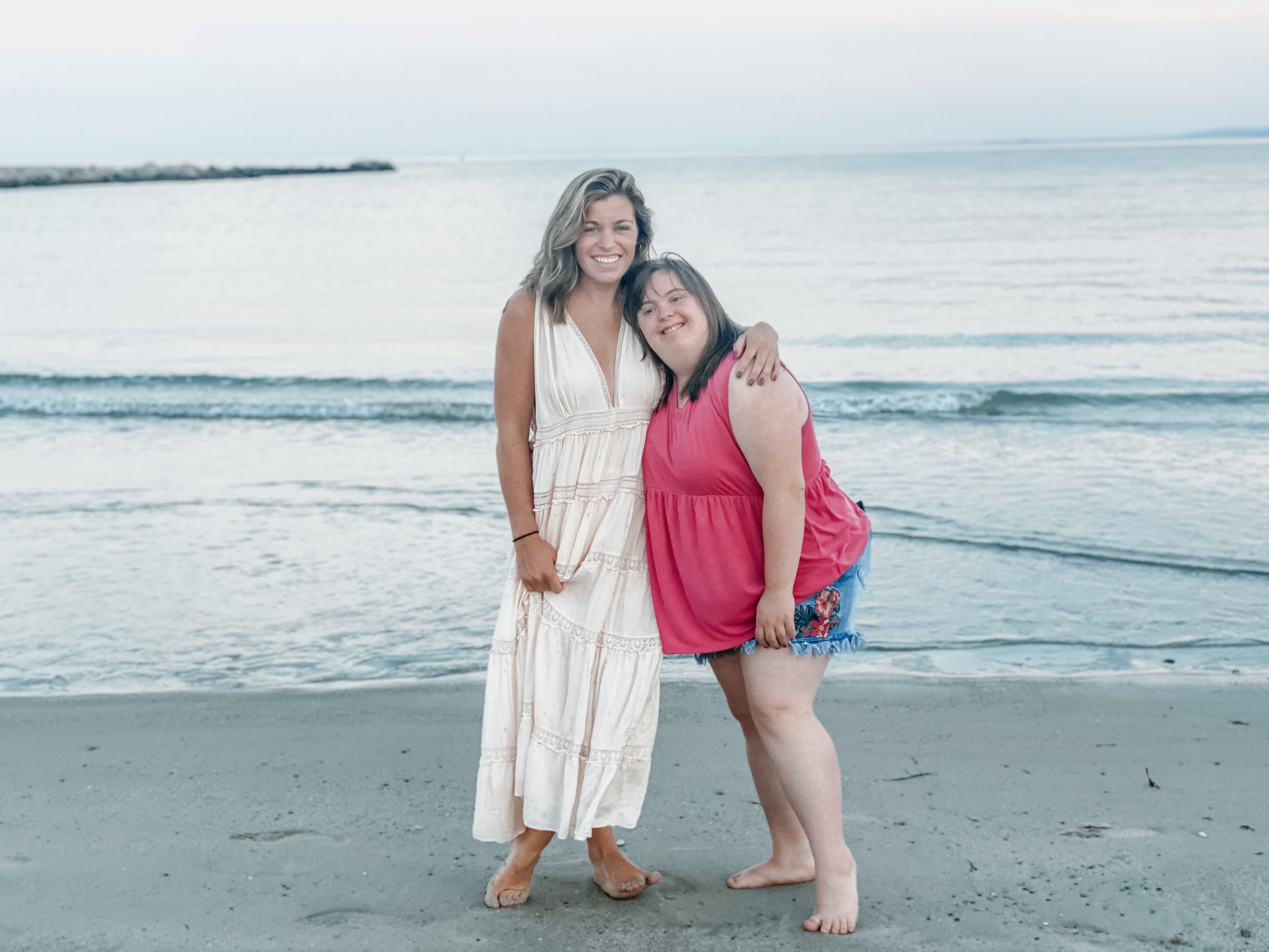 Two women standing on the beach near the water, smiling at the camera, wearing summer dresses and barefoot.