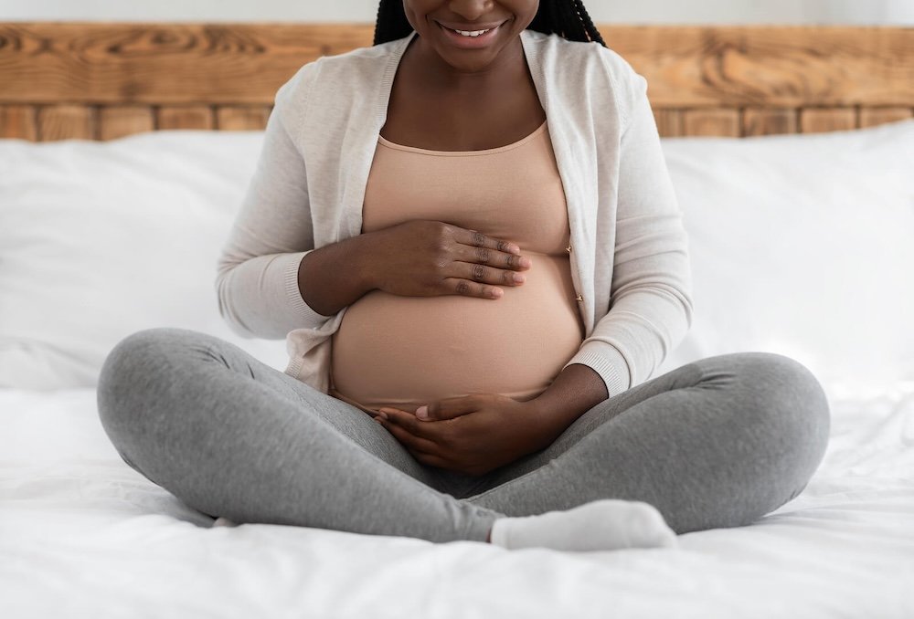 A pregnant woman sitting cross-legged on a bed, smiling, with her hands resting on her belly.