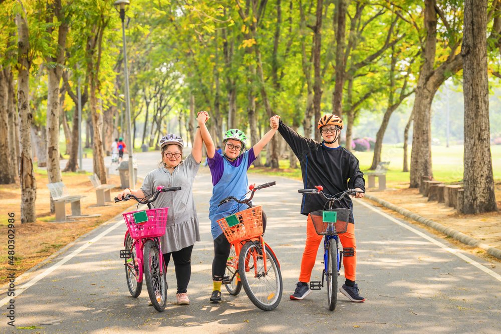 Three children wearing helmets holding hands while standing with their bikes on a park trail, trees lining the path on a sunny day.