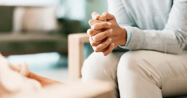 Person sitting with hands clasped, possibly in a thoughtful or prayerful pose, in an indoor setting.