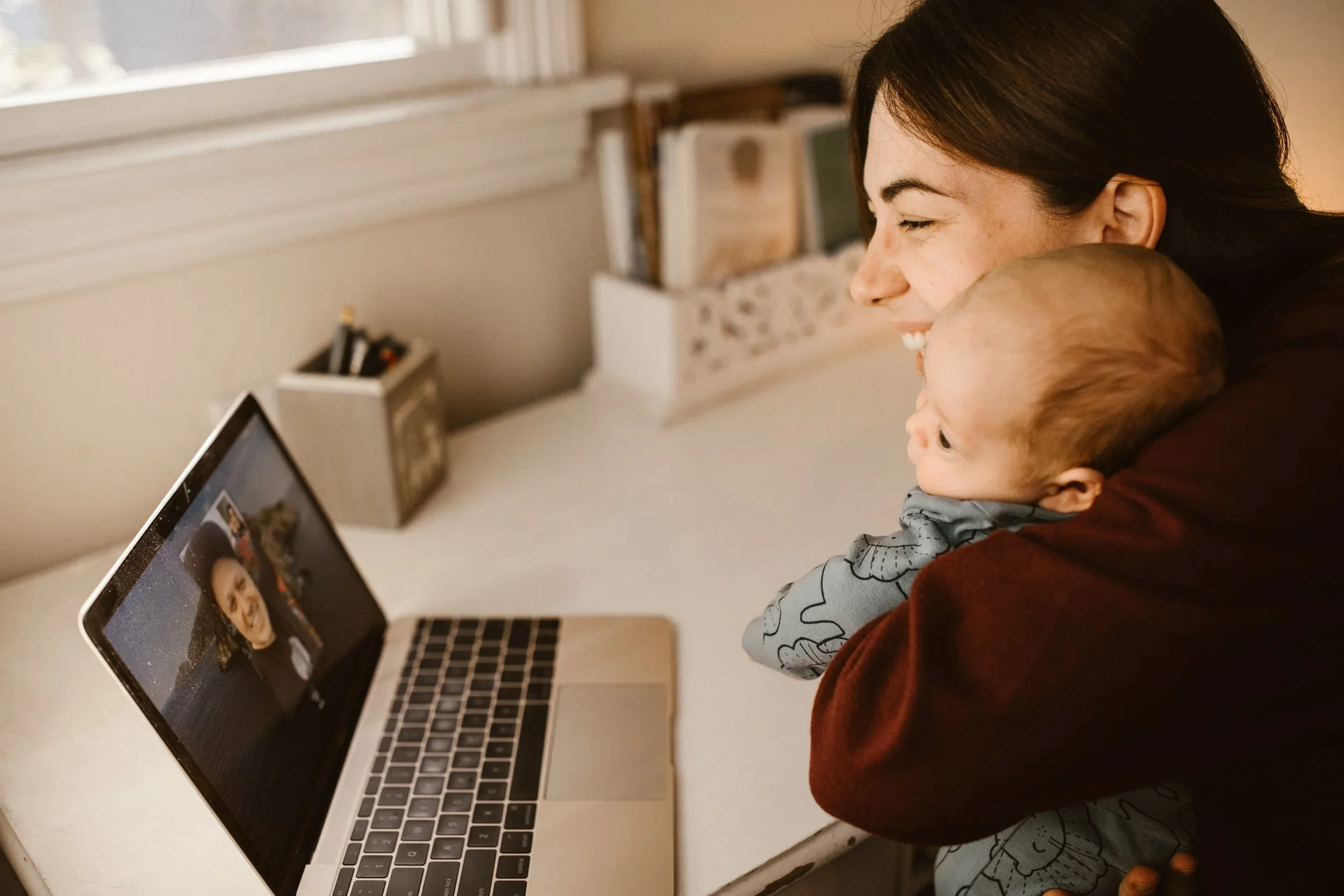 A woman and a young child watching a video call on a laptop screen, smiling while sitting close together at a desk near a window.