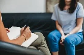 A person taking notes in a notebook during a therapy session with a woman sitting on a couch.