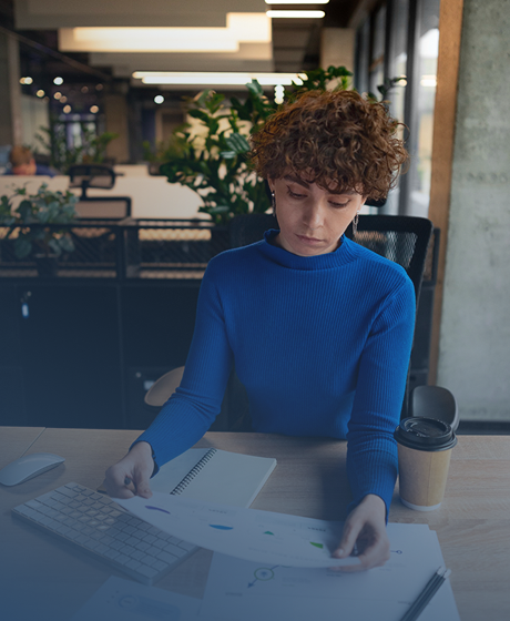Young woman with curly hair working at a desk with a keyboard, notebook, papers, and coffee in a modern office or cafe setting.
