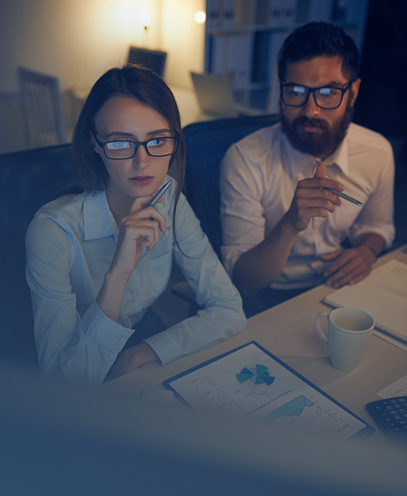 A man and woman wearing glasses working together at a desk with documents, coffee mug, and laptop in an office.