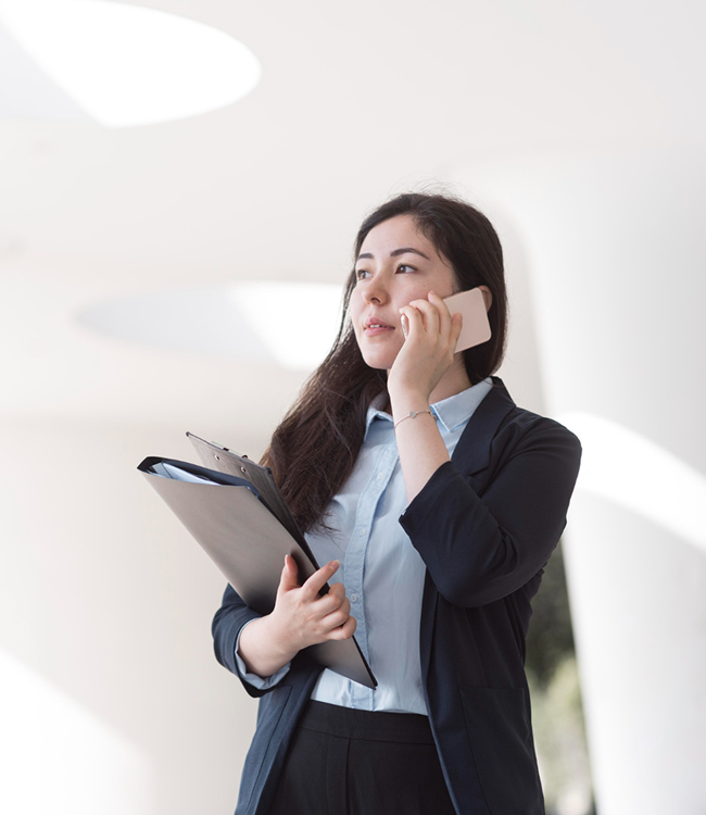 Businesswoman standing in a modern office, holding a clipboard and talking on a mobile phone.