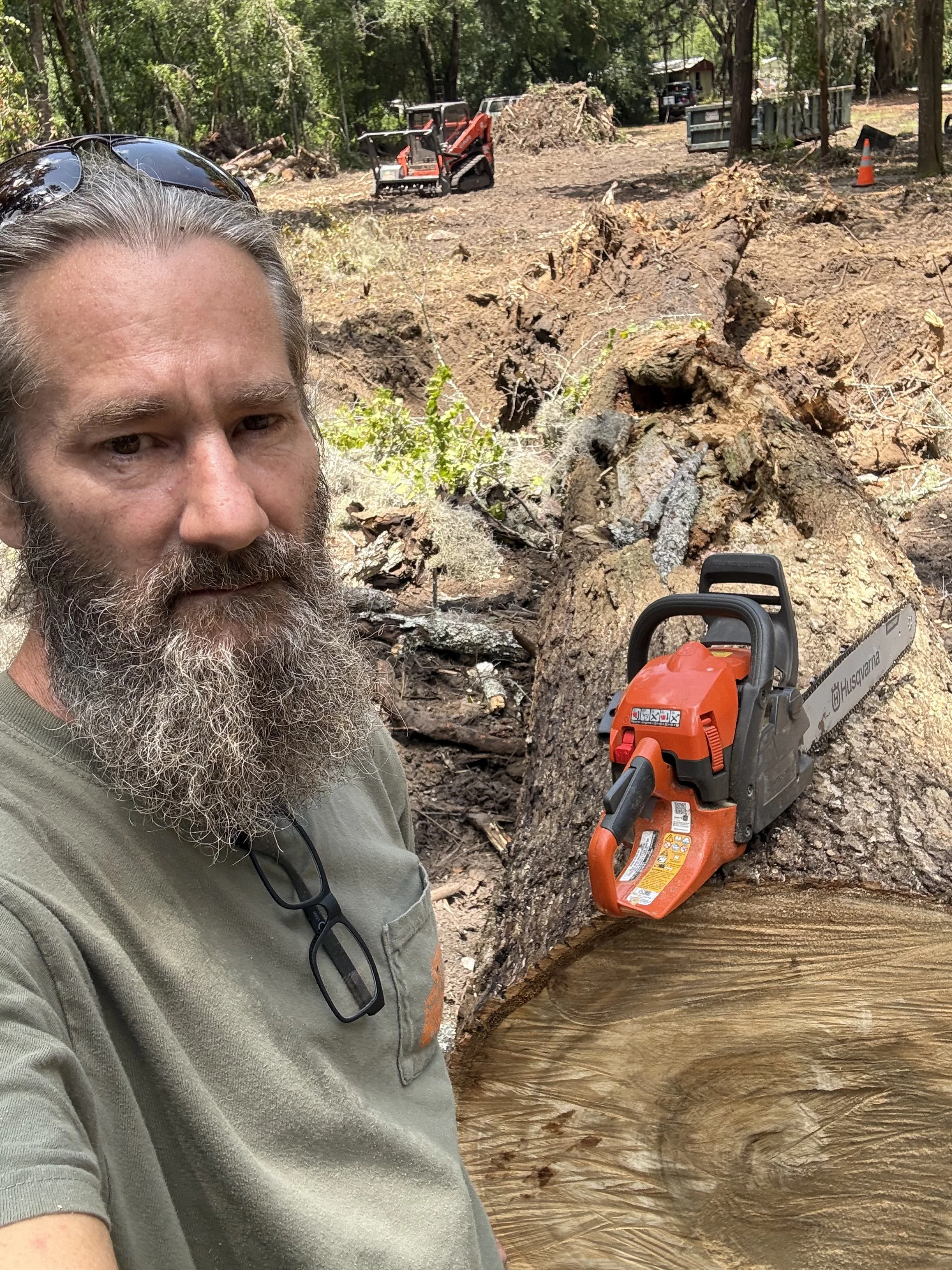 A man with sunglasses on his head, a beard, and a gray T-shirt stands in a wooded area. Behind him, a fallen tree is cut in half, with a chainsaw resting on it. In the background, there are trees, logs, and some equipment, including a small loader and traffic cones.