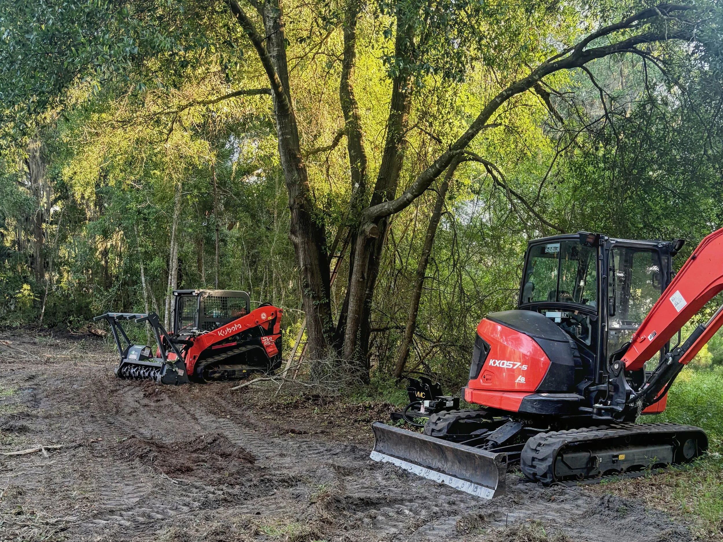 Construction equipment, including a mini excavator and rakes, parked on dirt near a wooded area with tall trees and dense greenery