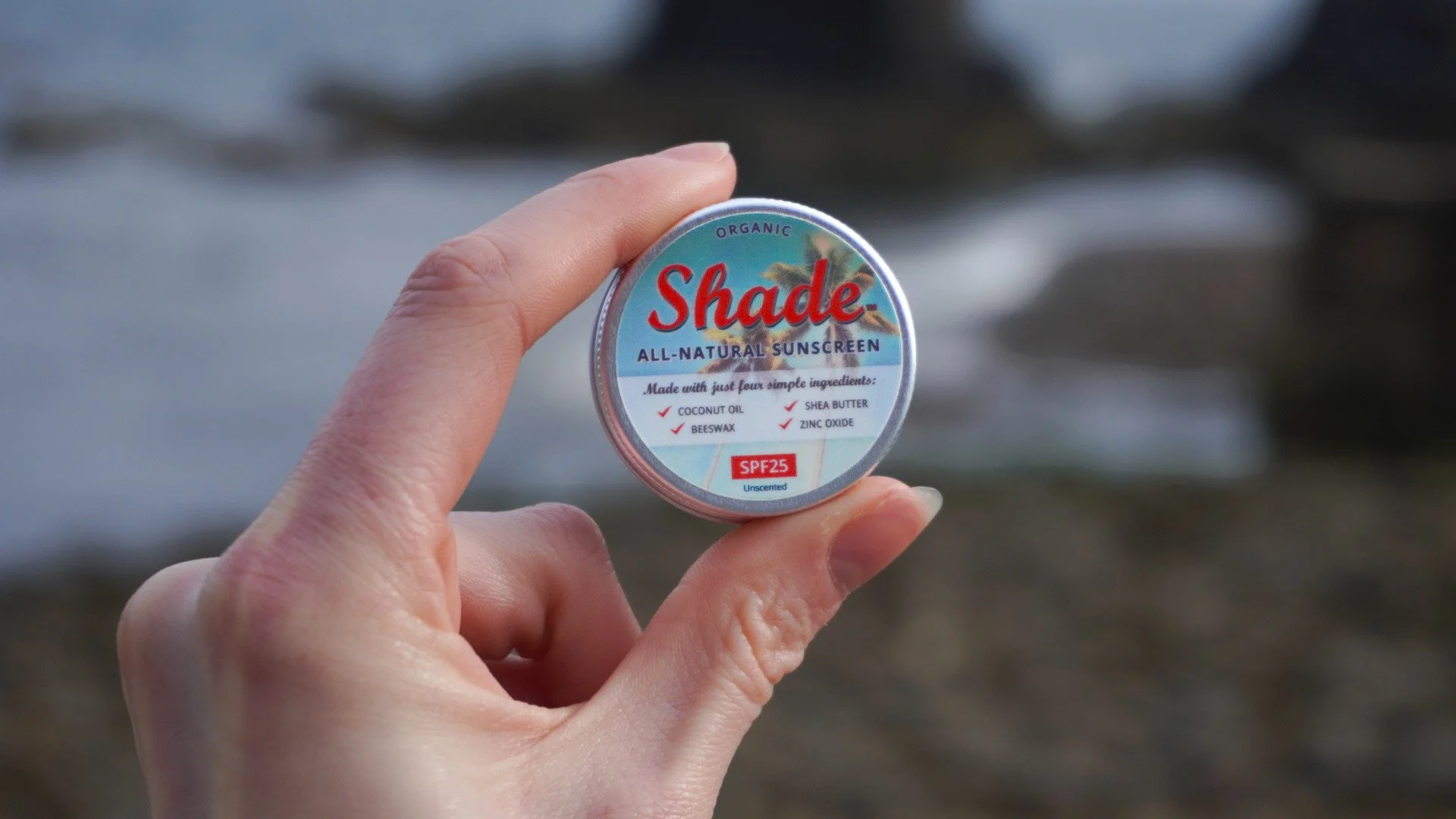 Minimalist brand imagery capturing a close-up of fingers holding a round tin of Shade all-natural organic sunscreen against a soft-focus beach backdrop in the Scottish Highlands.