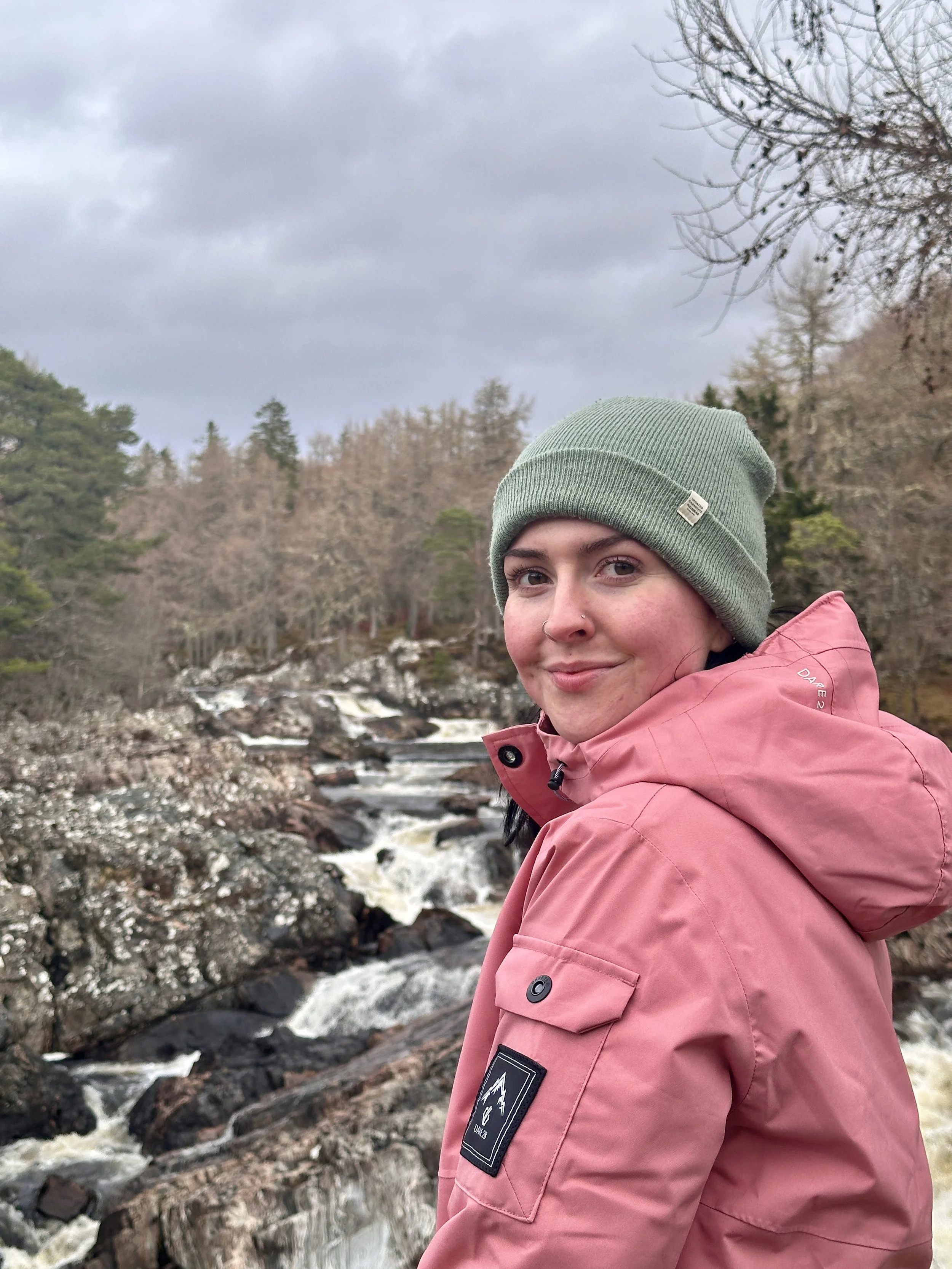 A woman with a nose piercing, wearing a sage green beanie and a pink ski jacket, standing in the Scottish Highlands by a rocky stream with a forest of trees in the background under a cloudy sky.