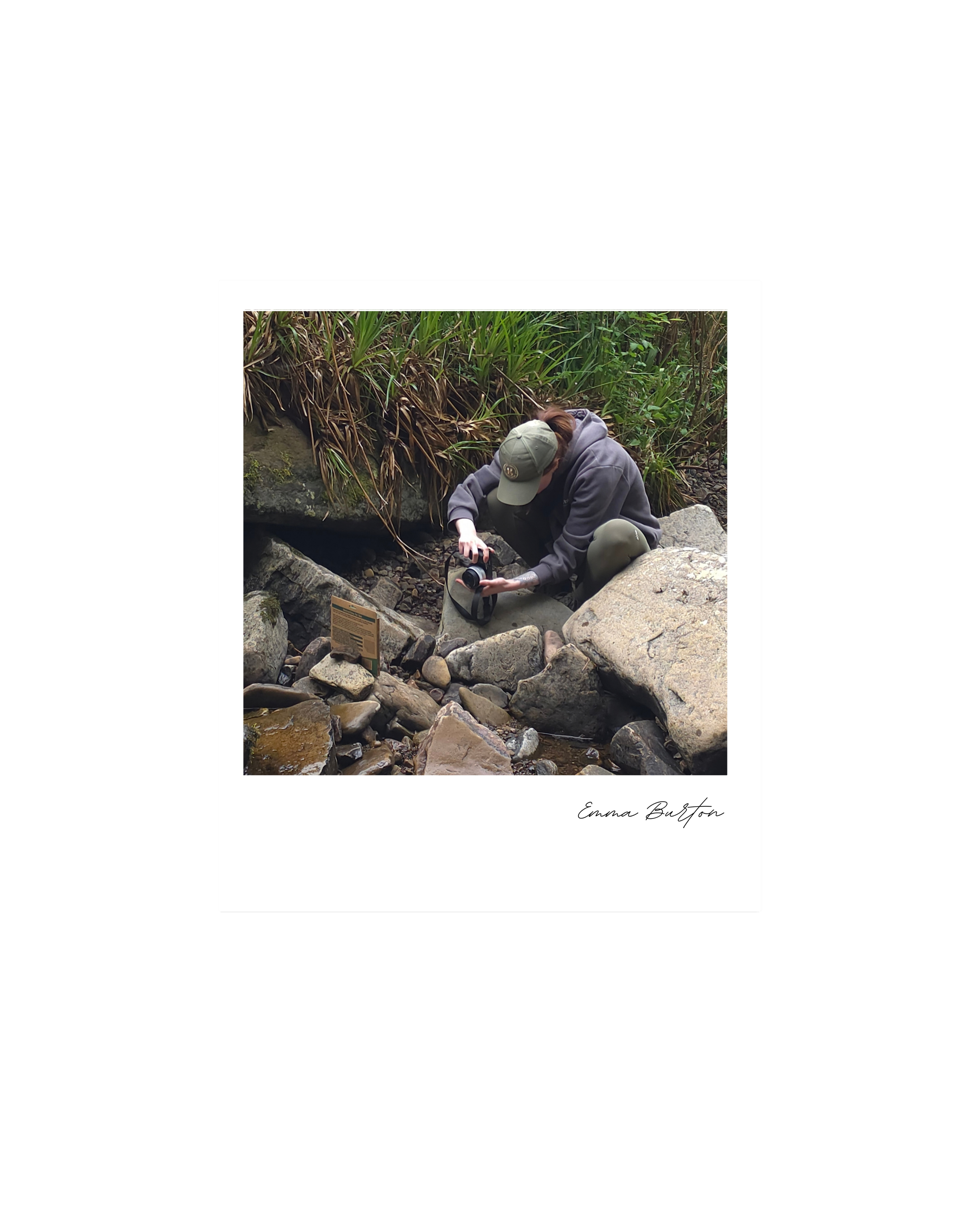 Emma Burton Studio crouches on the rocks by a stream in a lush, green forest. They are wearing a gray hoodie, gym leggings, and a green cap, and are holding a camera, taking product campaign pictures in the Scottish Highlands.