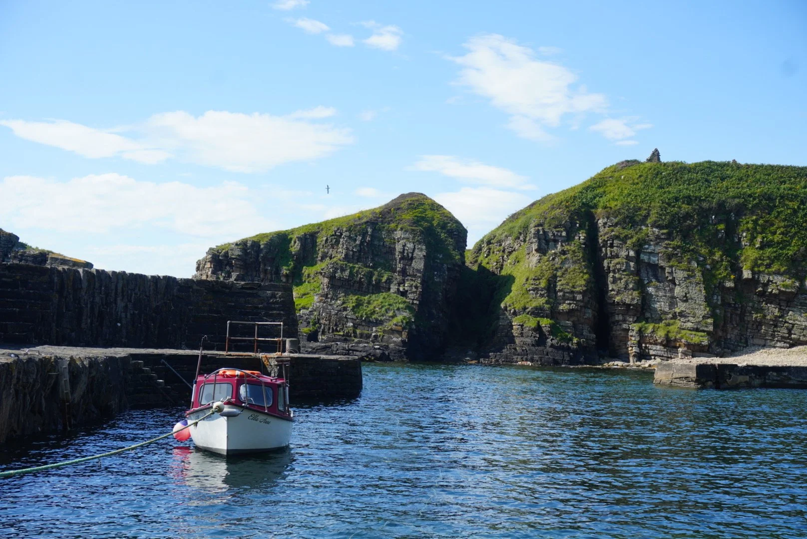 A small boat named Ella Jane floats in a Latheron Wheel harbor with dark stone walls. Behind the boat, there are large, rugged cliffs with green vegetation under a blue cloud sky in the Scottish Highlands, shot by Emma Burton Studio.