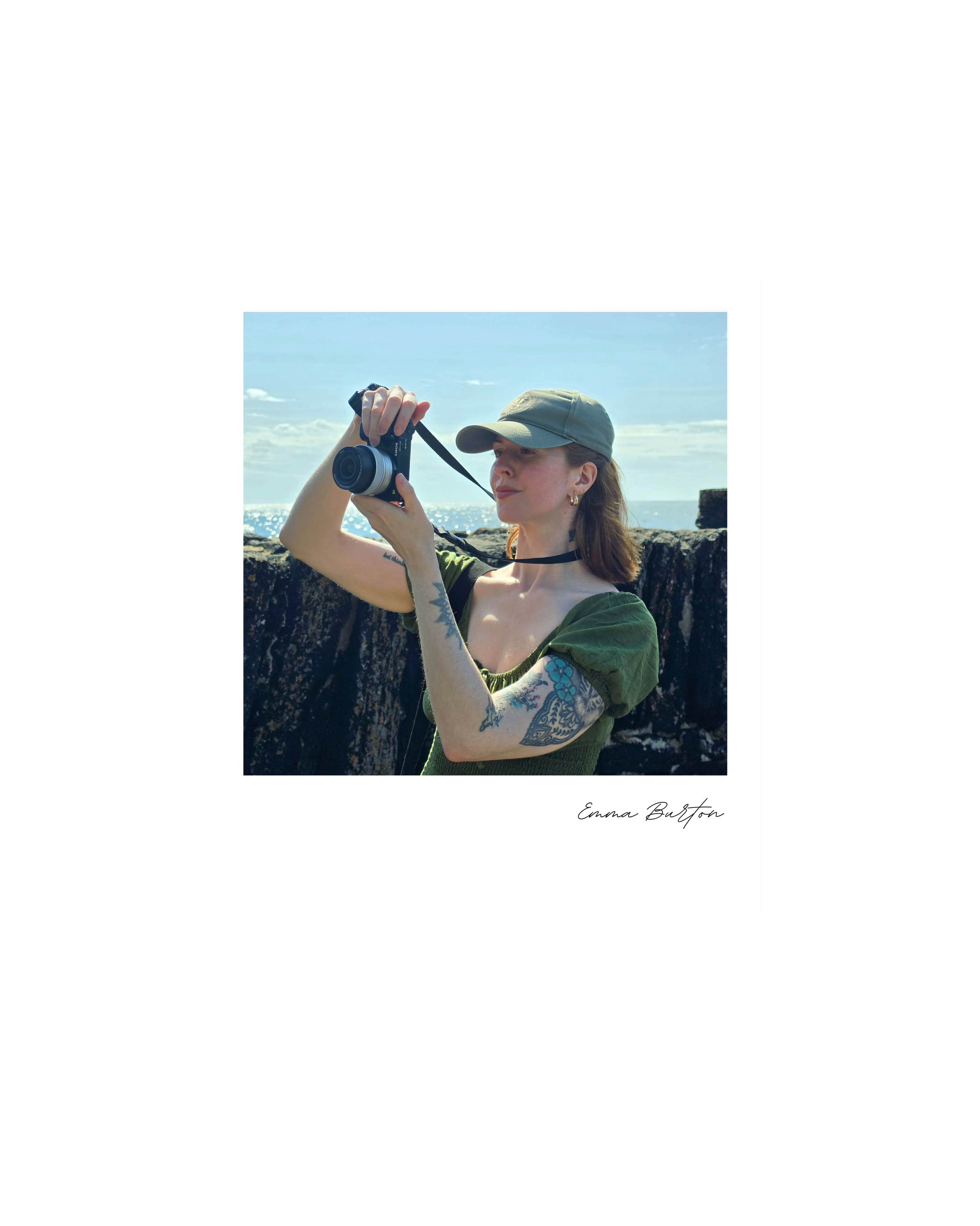 A woman with tattoos on her arm wearing a green shirt and a green cap taking campaign photos in the Scottish Highlands with a camera on a sunny beach with rocks and ocean in the background.