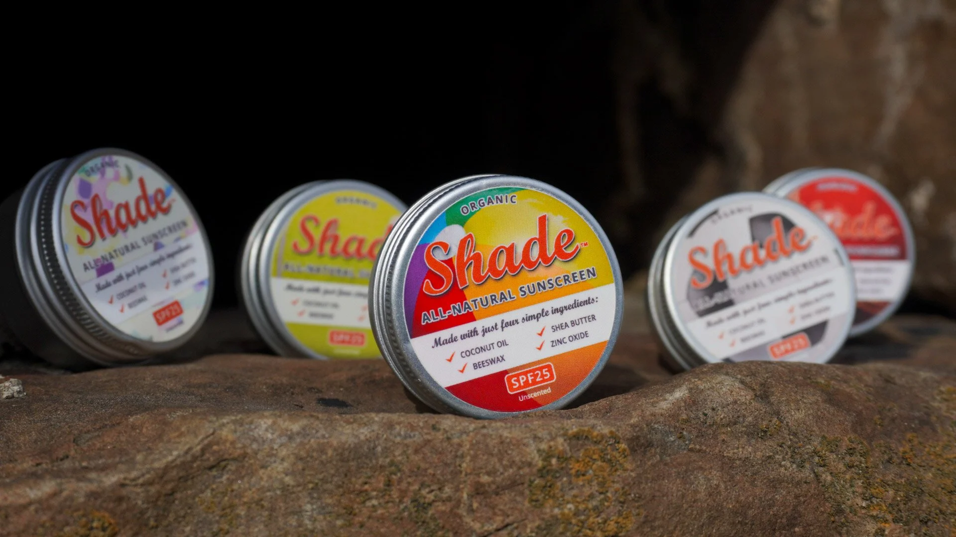 Professional commercial product photography displaying a range of Shade all-natural sunscreen tins arranged on a textured rocky outcrop in the Scottish Highlands. 