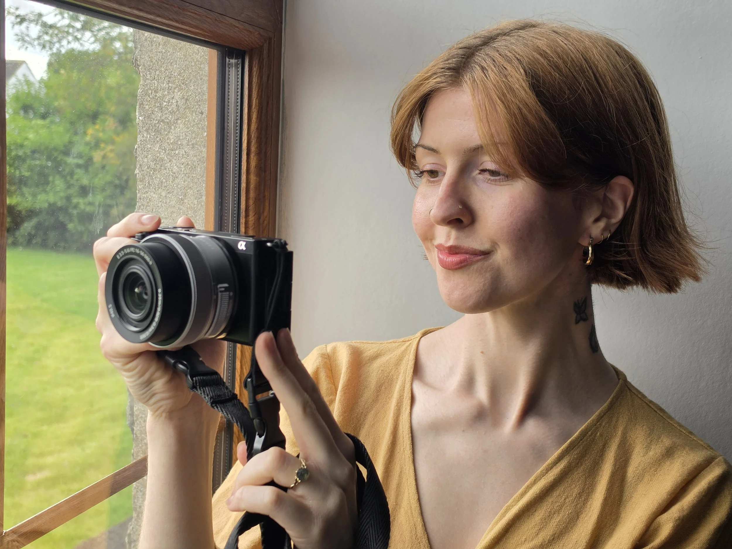A woman with auburn hair, a nose ring, and earrings taking a selfie with a Sony ZV-E10 camera near a window with a wooden frame, showing green trees outside shot by Emma Burton Studio.
