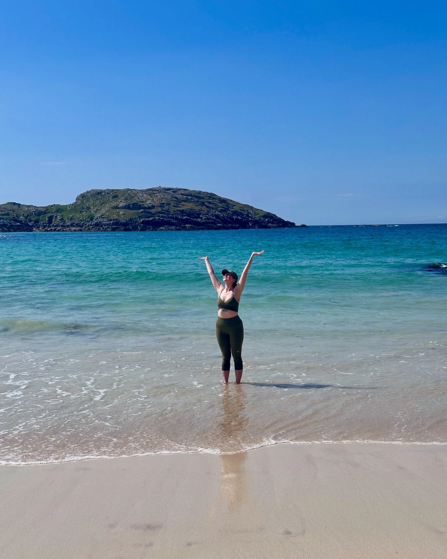 A woman standing in knee-deep blue water at the beach with her arms raised, wearing a green sports bra, matching leggings, sunglasses, and a cap, with an island and blue sky in the background on the west coast of the Scottish Highlands.